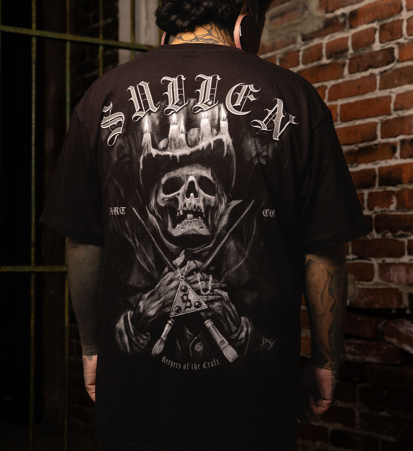 Person wearing a black t-shirt with a graphic design and text, standing against a brick wall.